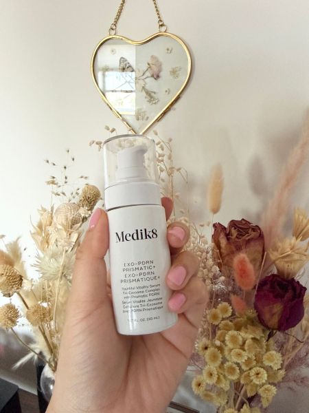 A hand holding the Medik8 ExoPDRN Prismatic serum in front of dried flowers